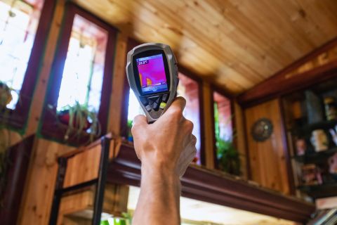A hand holding a thermal imaging camera scanning a wood-paneled ceiling for heat signatures