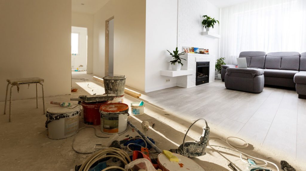 Paint buckets, dust, and renovation debris in a room with a finished living room visible beyond