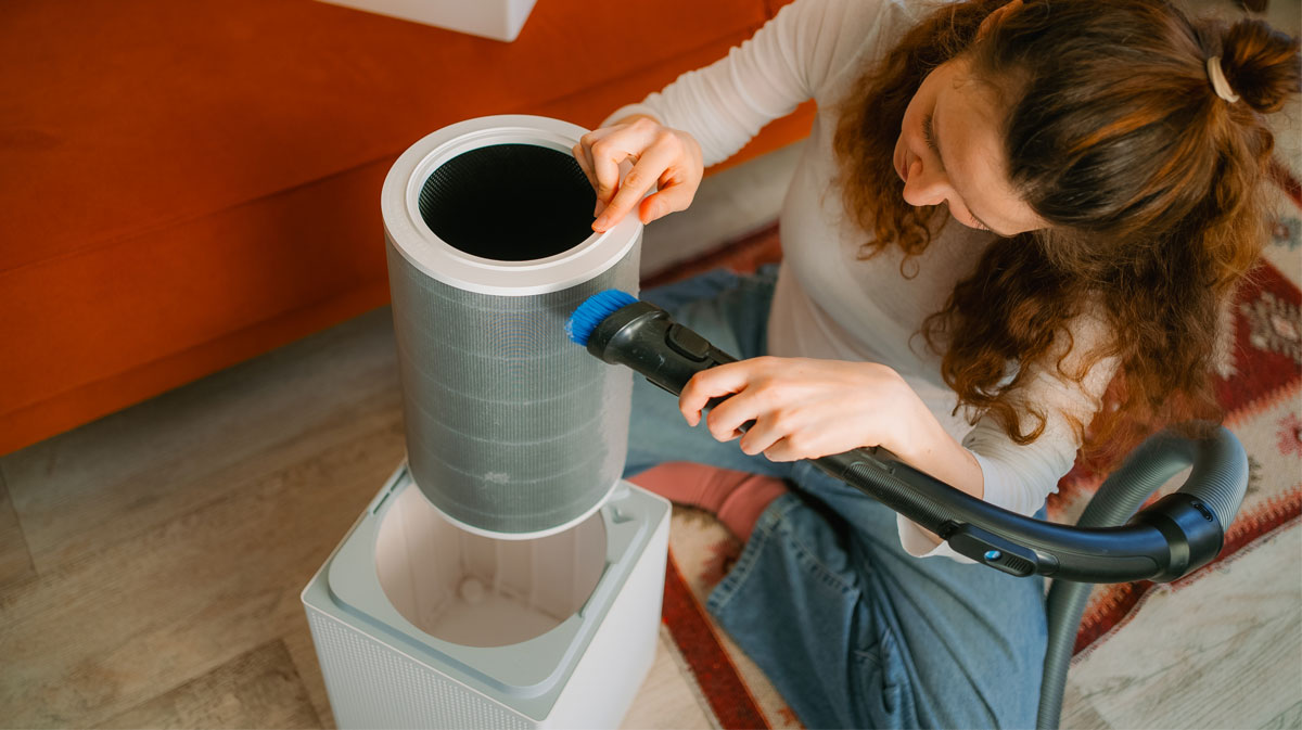 Woman vacuuming dust off a cylindrical HEPA air purifier filter with a brush attachment